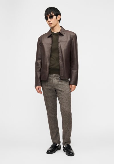 Άνδρας που φοράει σκούρο καφέ δερμάτινο μπουφάν, πουλόβερ σε ελιά πράσινο χρώμα, γκρίζο υφασμάτινο παντελόνι, μαύρα δερμάτινα μοκασίνια και μαύρα γυαλιά ηλίου, στέκεται μπροστά σε λευκό φόντο.