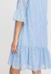 Rochie bleu deschis cu un tiv evazat, prezentând un model texturat în valuri și un material translucid. Mânici scurte cu volănașe accentuează designul său fluid.