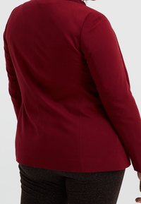 Burgundy tailored blazer with a smooth finish, featuring structured shoulders, a fitted silhouette, and minimal stitching details on the back.