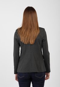 Blazer su misura grigio scuro con un design aderente, maniche lunghe, cuciture posteriori e una superficie liscia. Indossato sopra jeans blu scuro.