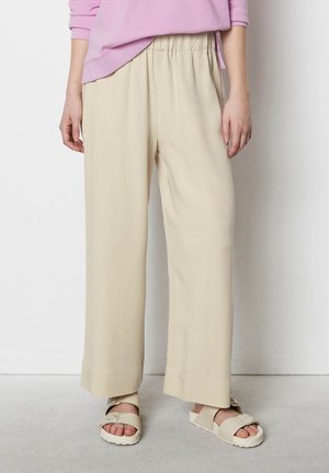 Person wearing loose beige pants, cream-colored buckle sandals, and a pink long-sleeve top standing against a white wall.