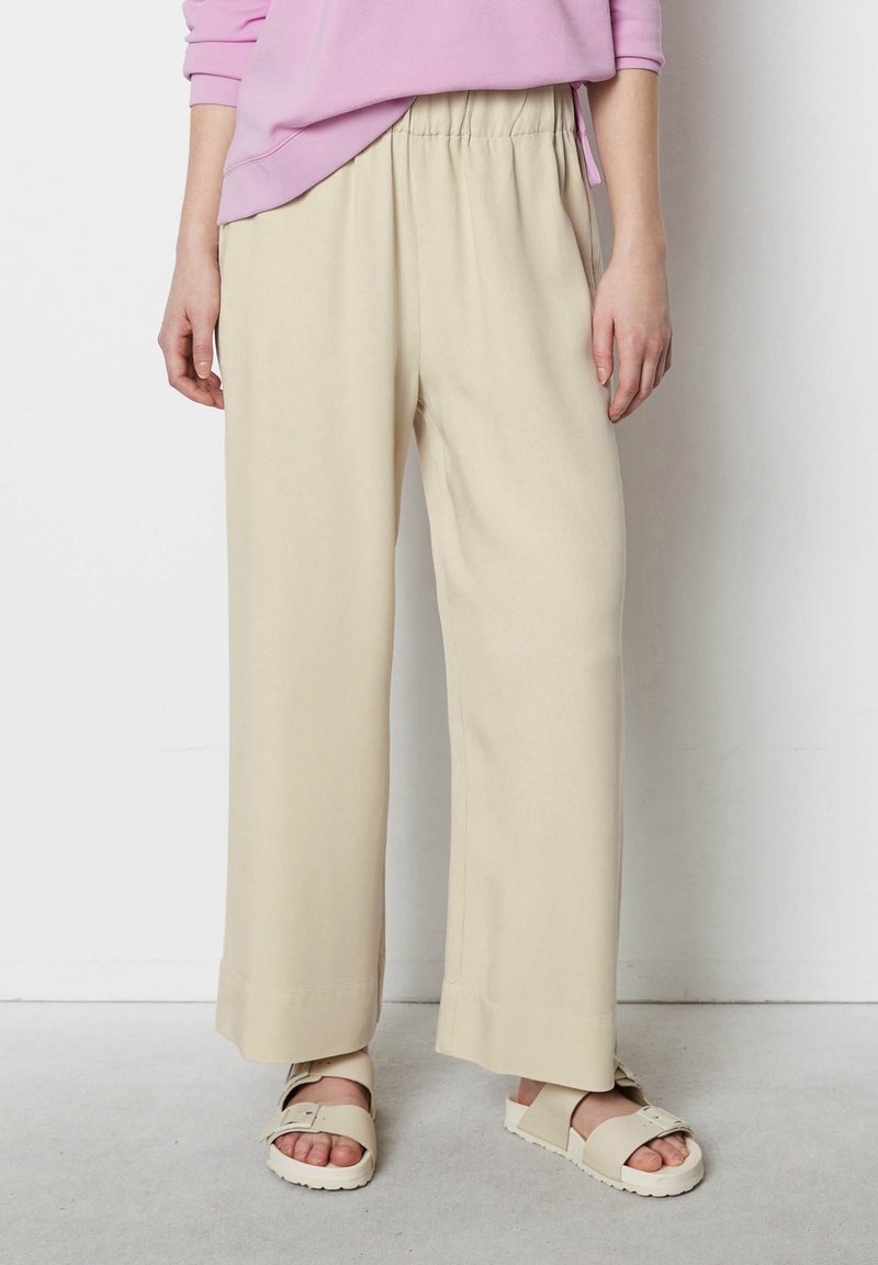 Personne portant un pantalon beige ample, des sandales à boucle couleur crème, et un haut rose à manches longues, debout contre un mur blanc.