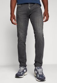 Jeans in denim grigio scuro slim-fit con una texture liscia, caratterizzati da un tradizionale design a cinque tasche e sottili dettagli di cucitura. Indossati con sneakers grigie.