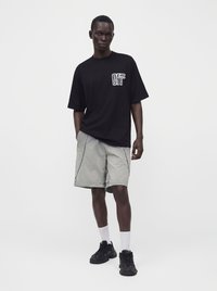 Άνδρας που φοράει μαύρο oversize μπλουζάκι με λευκό λογότυπο, γκρι σορτς με μαύρες ραφές, λευκές κάλτσες και μαύρα αθλητικά παπούτσια, στέκεται μπροστά σε λευκό φόντο.