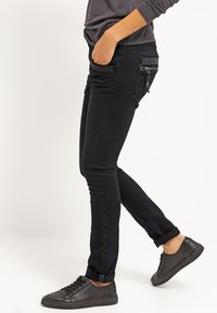 Černé úzké džíny s hladkým denimovým povrchem, zdobené dekoračním šitím a zipovými kapsami. V kombinaci s černými nízkými teniskami.