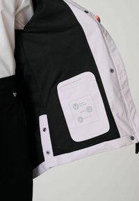 Interiér čierno-bielej bundy s sieťovou textúrou, ktorý obsahuje vrecúško s ikonami naznačujúcimi odolnosť voči dažďu, trvanlivosť a reflexnosť.