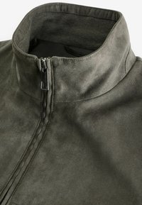 Veste en daim vert foncé avec un col haut et une fermeture éclair sur le devant. Texture lisse avec un léger éclat et des détails de couture soignés.