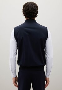 Marineblaues ärmelloses Oberteil aus glattem Stoff, mit hohem Kragen und geripptem Saum, kombiniert mit einem weißen Langarmshirt darunter.