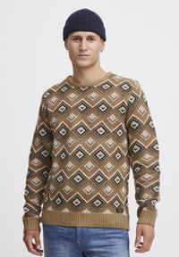 Bruine gebreide sweater met een patroon van diamanten in crème, zwart en groen, met geribde manchetten en zoom, gecombineerd met blauwe spijkerbroek.