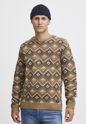 Brown patterned sweater featuring diamond shapes in cream, black, and green, with ribbed cuffs and hem, paired with blue jeans.