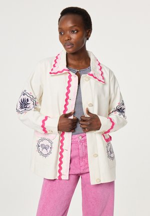 Woman wearing a white jacket with pink trim and embroidered sleeves over a striped top and pink pants, standing against a plain background.