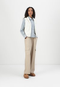 Gilet en tricot blanc sur une chemise boutonnée rayée bleue et blanche, associé à un pantalon cargo beige avec des poches latérales et des chaussures à plateforme marron.