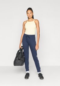 Ανοιχτό κίτρινο αμάνικο τοπ, σκούρα μπλε φόρμα με λευκές λεπτομέρειες λογότυπου, μαύρη γυμναστηριακή τσάντα και μαύρα αθλητικά παπούτσια σε ουδέτερο φόντο.