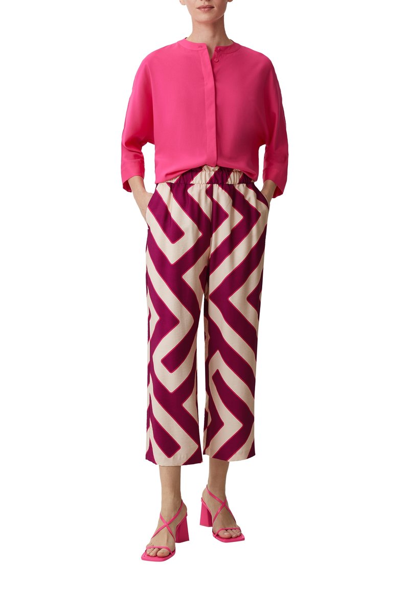 Kobieta ubrana w jasnoróżową bluzkę, kremowe i purpurowe szerokie spodnie w geometryczne wzory oraz różowe sandały na obcasie z paskami.