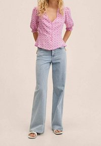 Blouse rose à motifs avec manches bouffantes, associée à un jean évasé bleu clair et des sandales blanches à bouts ouverts. Détails du tissu texturé visibles.