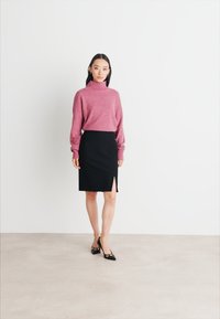 Pulover roz cu gât de broască, asortat cu o fustă creion neagră cu o deschidere pe lateral. Modelul poartă tocuri negre cu accente aurii. Fond simplu.