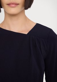 Haut bleu marine avec un décolleté carré, des plis sur l'épaule et un tissu lisse et extensible. Le design met en avant la simplicité et l'élégance.