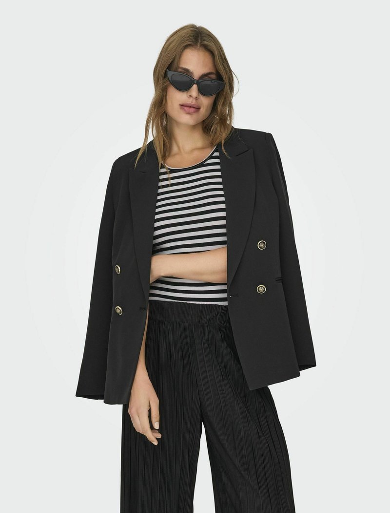 Zwarte dubbelgeknoopte blazer met gouden knopen, gedragen over een cropped zwart-wit gestreept topje. Plissé zwarte broek met wijde pijpen en donkere zonnebril.