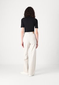 Femme aux cheveux bouclés vue de dos, portant un haut noir à manches courtes, un pantalon blanc large taille haute et des baskets blanches sur fond blanc.