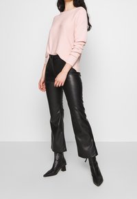 Femme portant un pull à manches longues rose clair, un pantalon taille haute en similicuir noir et des bottines à talons pointues noires.