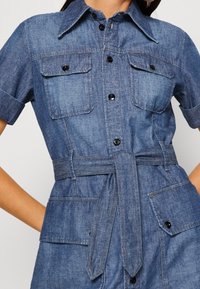 Jeansklänning med korta ärmar, frontknäppning, två bröstfickor och ett bälte i tyg vid midjan. Medelblå färg.