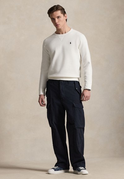 Mladi moški, oblečen v belo majico z dolgimi rokavi, temne cargo hlače in superge, stoji pred enobarvnim bež ozadjem.