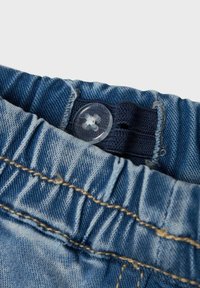 Denim püksid, millel on kogutud elastne vöökoht, selgeplastikust nööp ja kontrastsed sinised õmblused.