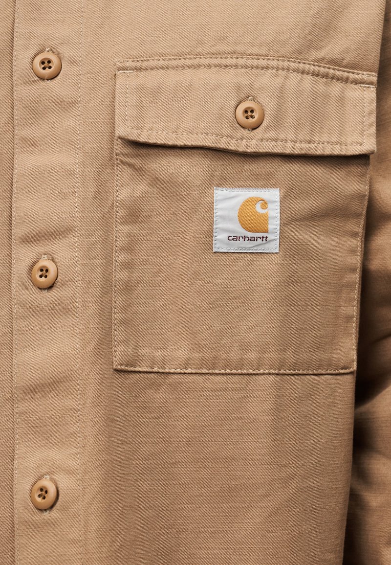 Camicia abbottonata color sabbia, tessuta, con taschino sul petto, patch con logo rotondo e bottoni marroni. Tessuto strutturato con una finitura liscia.