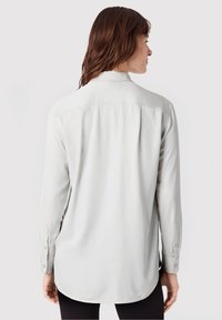 Eine Frau mit braunen Haaren trägt ein langärmliges, hellgraues Hemd mit Knöpfen, von hinten gesehen, vor einem schlichten Hintergrund.