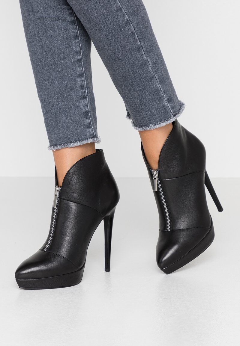 Bottes noires à talons hauts avec fermetures éclair à l'avant, portées avec un jean gris à ourlet effiloché sur un sol blanc.