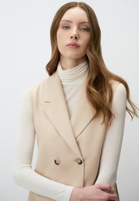 Gilet beige doppiopetto con rever a punte, realizzato in tessuto liscio, abbinato a un dolcevita cremoso a costine. Capelli lunghi e mossi.