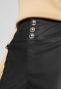 Zwarte leren broek met een hoge taille en drie met skulls versierde gouden knopen. De stof heeft een gladde textuur met zichtbare stiksels.