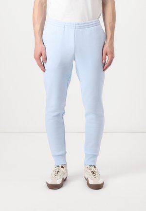 Mann trägt hellblaue, enge Jogginghosen und weiße Turnschuhe und steht vor einem schlichten weißen Hintergrund.