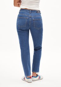Blå denimjeans med rak benpassform, försedda med två bakfickor, sömnadsdetaljer och en midjebandsetikett synlig på baksidan.