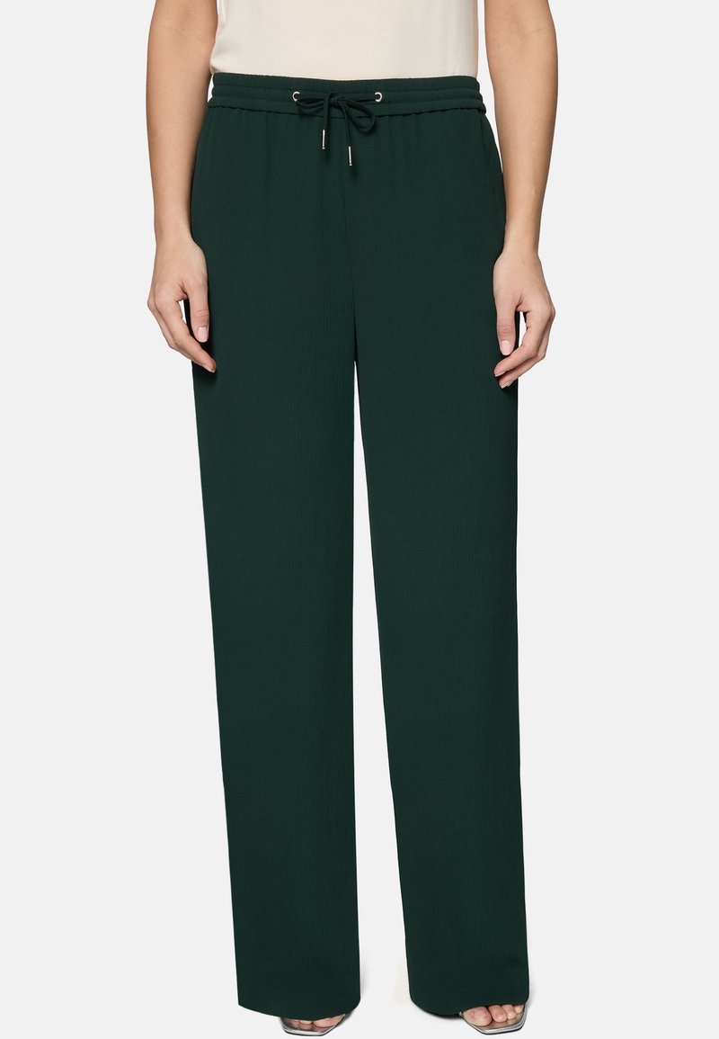 Pantalon large vert foncé à taille coulissante porté par une personne debout devant un fond uni.