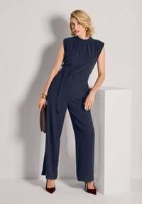 Vrouw in een marineblauwe mouwloze jumpsuit met tailleband, bordeaux hakken, die een textuur clutch vasthoudt en leunt op een witte rechthoekige voetsteun.