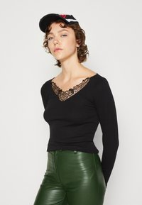 Svart langermet topp med blond V-hals, tettsittende design, sammen med glinsende grønne faux skinnbukser.