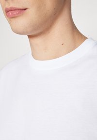White cotton t-shirt with a crew neckline, smooth texture, and short sleeves, showing part of a neck and jawline.