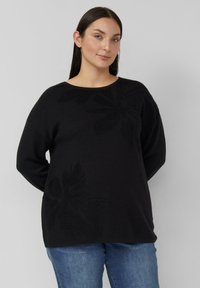 Frau mit langen dunklen Haaren, die einen schwarzen Pullover mit dezentem Blattmuster und blaue Jeans trägt, steht vor einem schlichten, hellen Hintergrund.