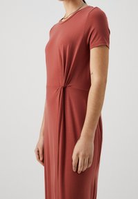 Sukienka ma kolor rdzy, wykonana jest z miękkiego, elastycznego materiału. Posiada okrągły dekolt i detal w postaci węzła w talii, zwężający się ku dołowi.