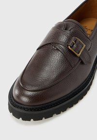 Mocassins en cuir marron à finition texturée, dotés d'une large lanière avec un détail de boucle en or. Semelle épaisse en caoutchouc noir avec un motif rainuré.