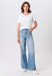 White short-sleeve shirt with a rounded neckline, paired with light blue wide-leg jeans featuring intricate floral embroidery on the pockets.