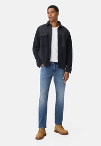 Marine fleece shirtjack met voorzakken, over een wit T-shirt. Lichtblauwe jeans en tan kleurige veterschoenen maken de outfit compleet.