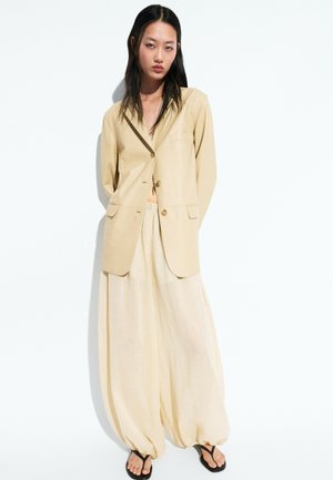 Jonge vrouw met lang donker haar die een beige blazer, losse beige broek en zwarte sandalen draagt tegen een witte achtergrond.