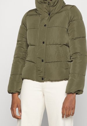 Personne portant une doudoune matelassée vert olive avec des boutons-pression et un pantalon blanc, debout devant un fond uni.