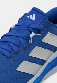 Sinine spordijalats, millel on tekstureeritud võrgusine pealispind, lamedad paelad ja kolm metallik-hõbedast triipu küljel. Kerge disain ja pehmenduse detailid.