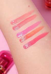 Fargeprøver av fem leppeprodukter på en lys arm. Fargene inkluderer myk rosa, sterk rosa, lilla, koralloransje og dyp rosa. Skinnende finish.