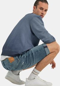 Sudadera azul de cuello redondo, pantalones cortos de mezclilla con puños doblados, zapatillas blancas y calcetines a rayas. Etiqueta de cuero destacada en los pantalones cortos.