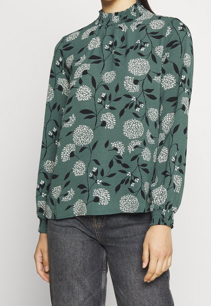 Green floral blouse with white and black patterns, gathered neckline, and elastic cuffs. Material appears lightweight with a smooth texture.