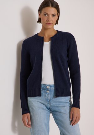 Femme aux cheveux courts et foncés porte un cardigan zippé en tricot bleu marine sur une chemise blanche et un jean taille haute bleu clair sur un fond uni.